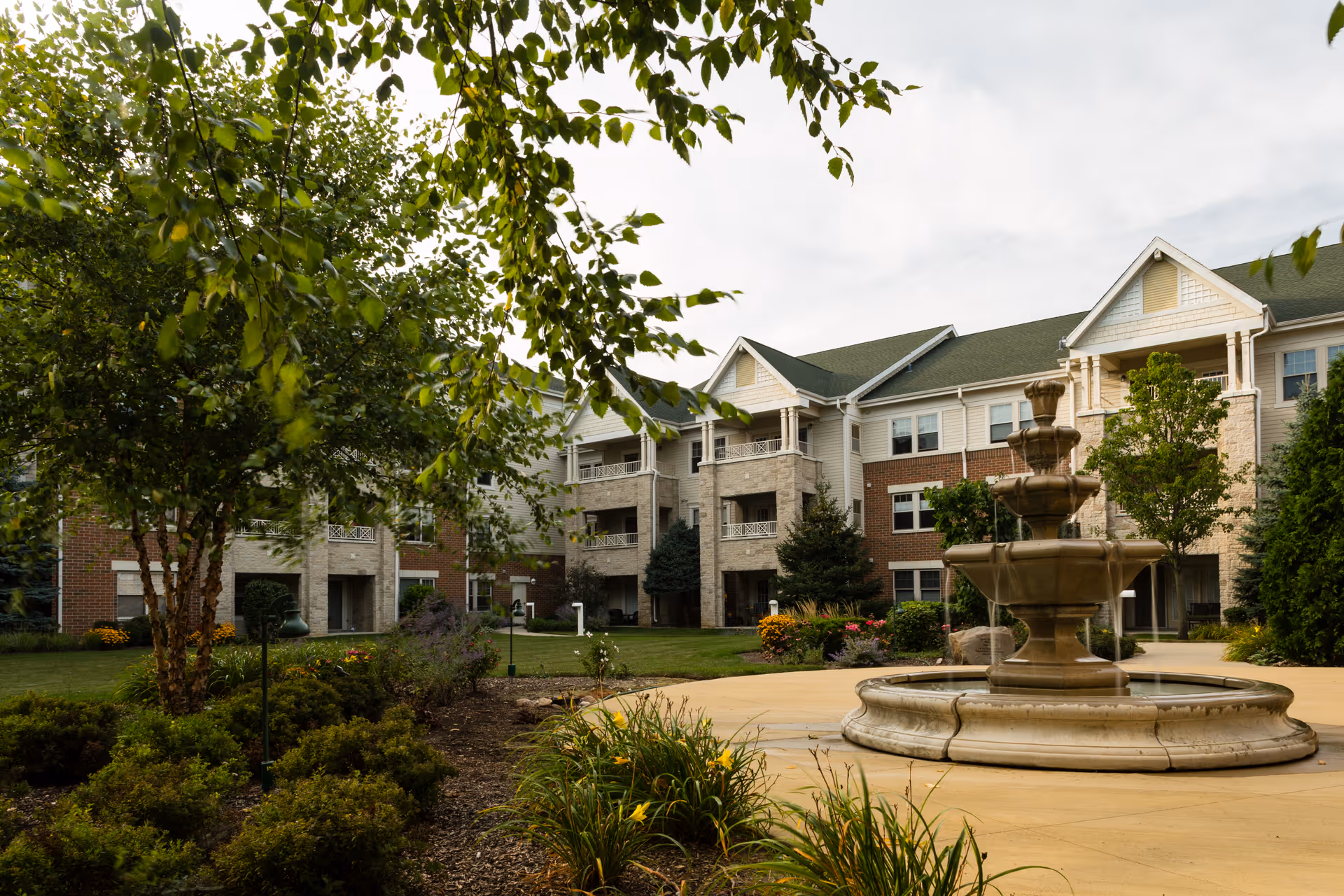 Courtyard with a multi-tier fountain and landscaped grounds in front of a three-story senior living building.