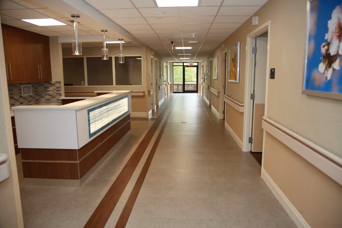 A clean and well-lit hallway in a senior living facility with a reception desk on the left side, beige walls with handrails, framed floral artwork on the right wall, and a glass door at the end of the corridor letting in natural light.