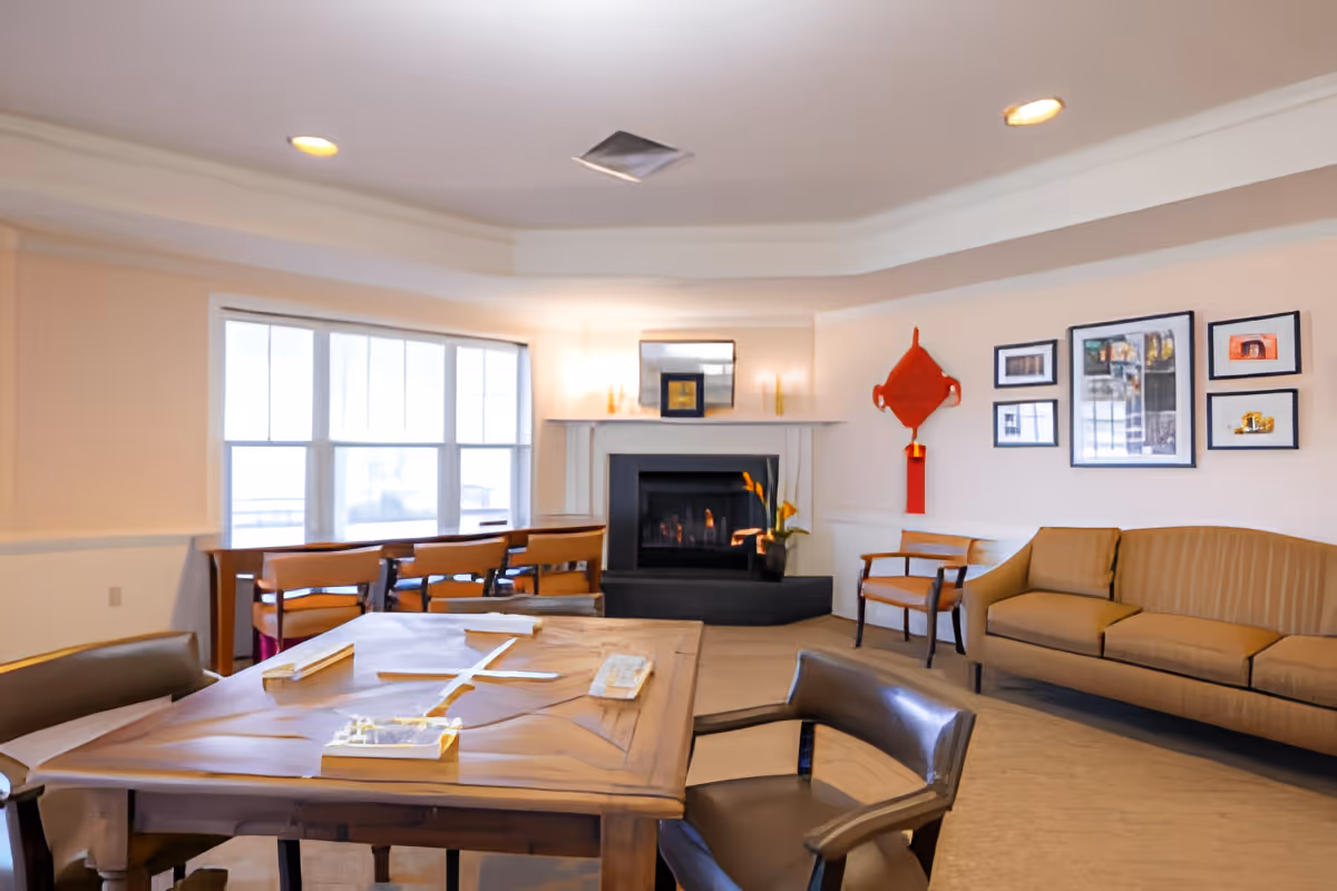 A cozy living room area with a wooden table and chairs in the foreground, a beige sofa and wooden armchairs along the walls, a fireplace with a fire burning, and a large window letting in natural light. The walls are decorated with framed pictures and a red hanging ornament.