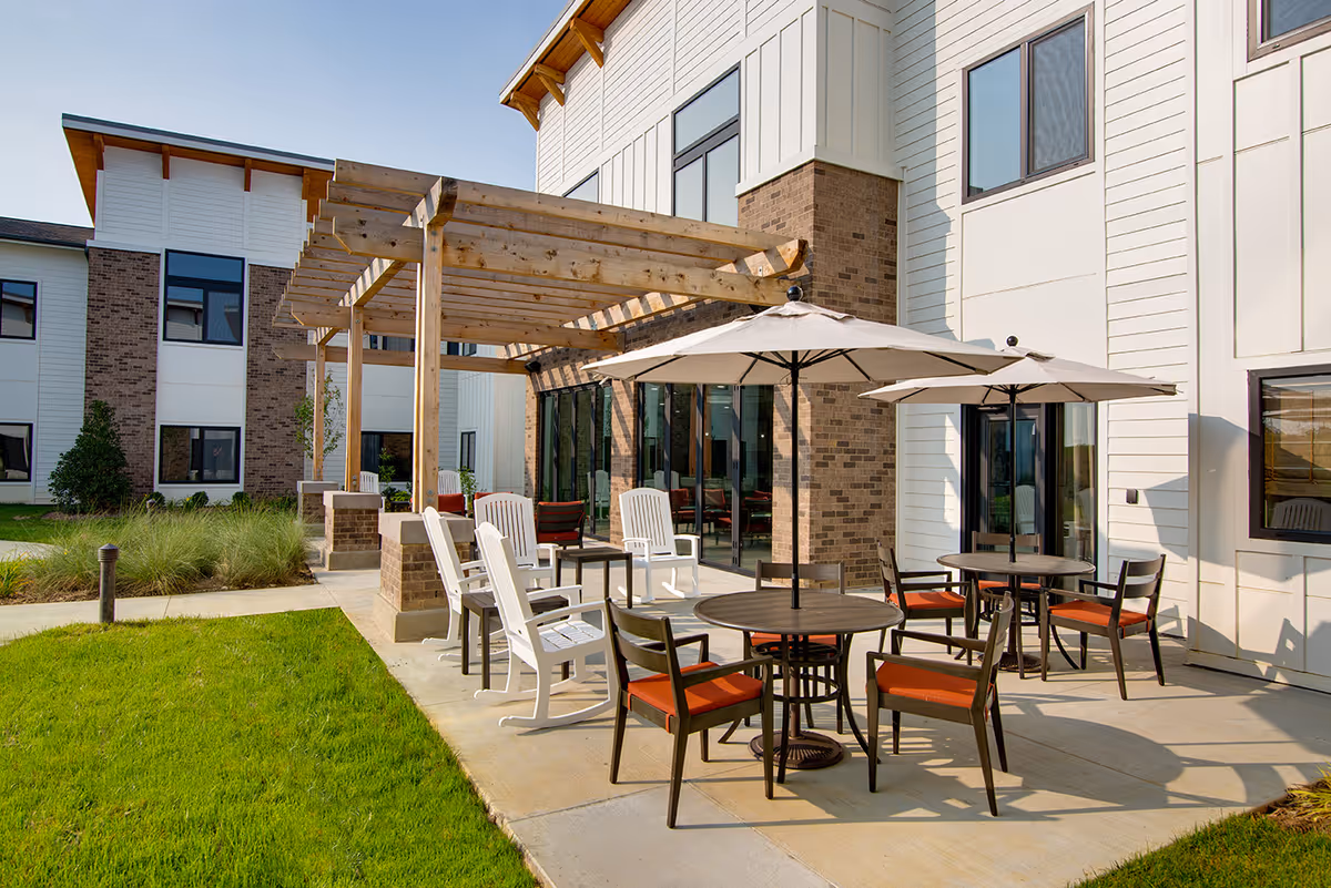 Outdoor patio at a senior living facility with round tables, umbrellas, chairs and a wooden pergola beside the building.