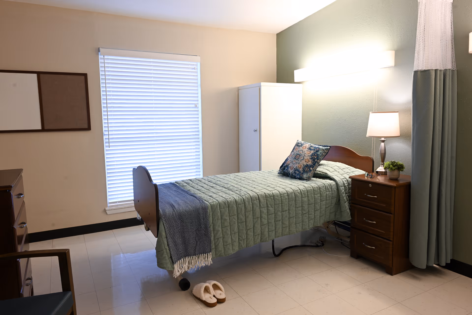 A neatly made single bed with a green quilt and a decorative pillow in a nursing facility room. Next to the bed is a wooden nightstand with a lamp and a small plant. There is a white wardrobe against the wall and a window with closed blinds. A pair of slippers is placed on the floor near the bed. The room has a calm and clean atmosphere with light-colored walls and tiled floor.