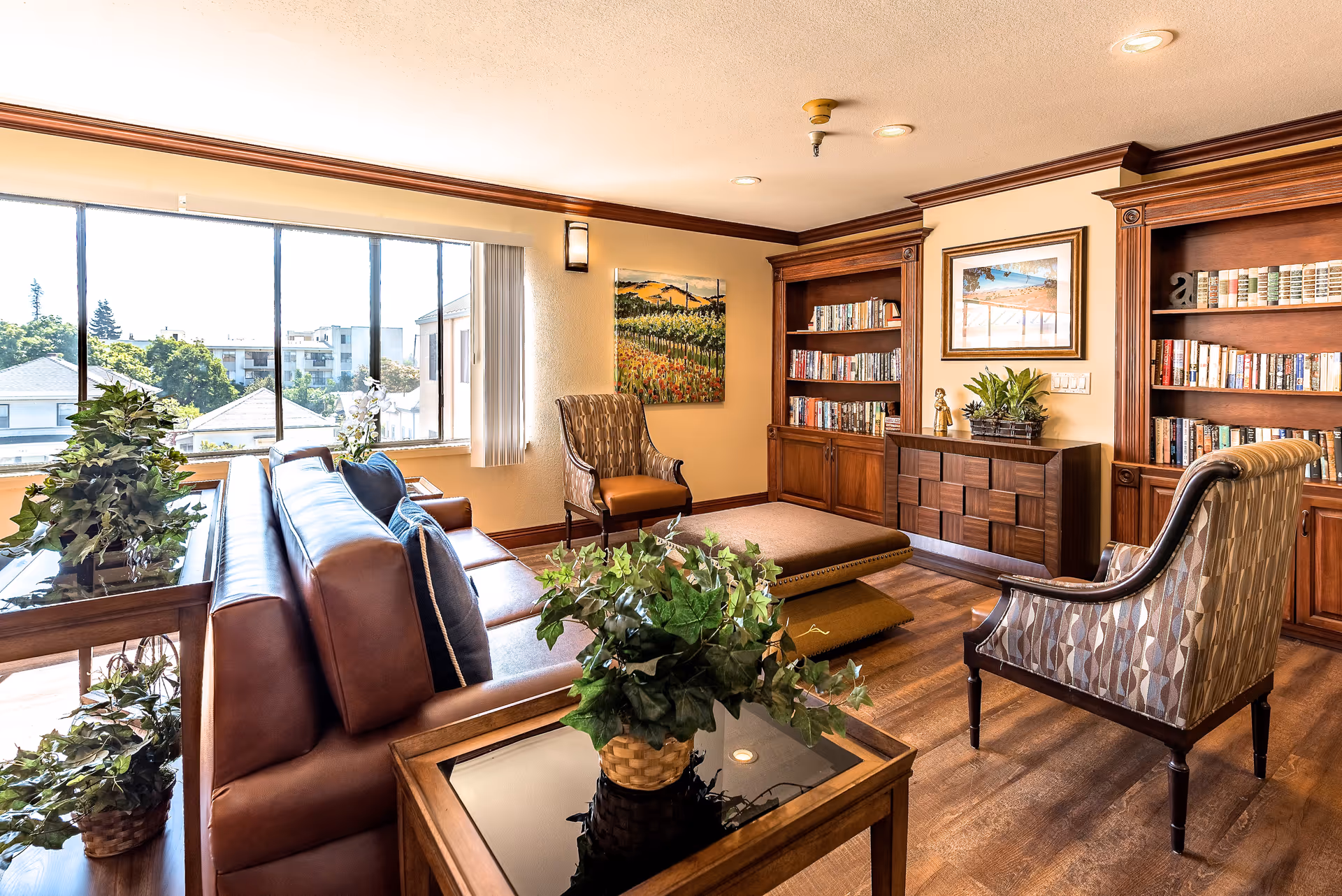 A cozy senior living room with large windows letting in natural light, a brown leather sofa with blue cushions, two patterned armchairs, a wooden coffee table with a potted plant, and built-in wooden bookshelves filled with books. The room has warm wood flooring and decorative paintings on the walls.