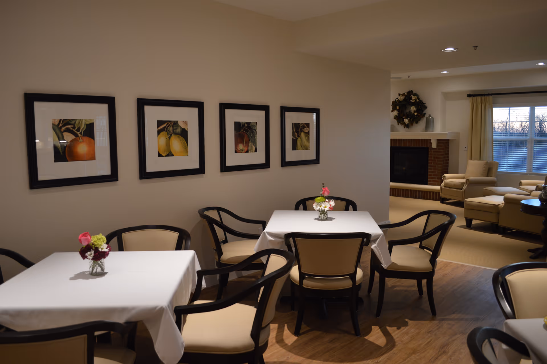 A cozy dining area with two tables covered in white tablecloths, each adorned with small floral centerpieces. The tables are surrounded by cushioned chairs with dark wooden frames. On the wall above the tables, there are four framed pictures of fruit. In the background, a living room area is visible with beige armchairs, a fireplace decorated with a wreath, and a window with curtains letting in natural light.