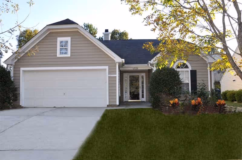 Front exterior view of a single-story house with beige siding, a two-car garage, a concrete driveway, and a small front yard with green grass and some shrubs and flowers near the entrance.
