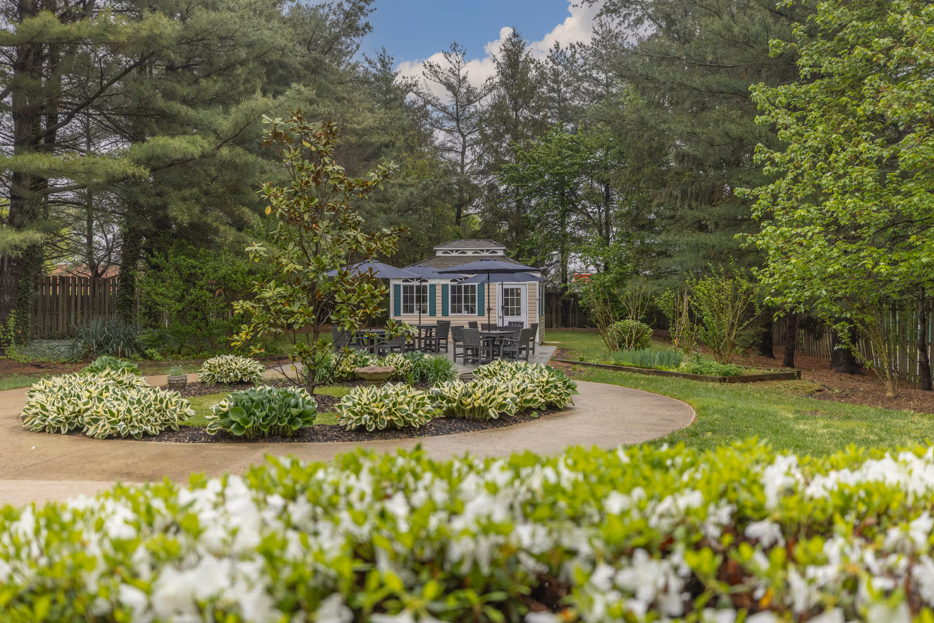 A landscaped outdoor garden area with a curved concrete pathway, surrounded by green shrubs and trees. In the background, there is a small building with blue shutters and a patio area with tables and chairs under large umbrellas.