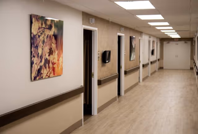 A long, well-lit hallway in a healthcare or rehabilitation facility with light wood flooring, beige and white walls, handrails on both sides, several open doorways, and framed floral artwork hanging on the walls.