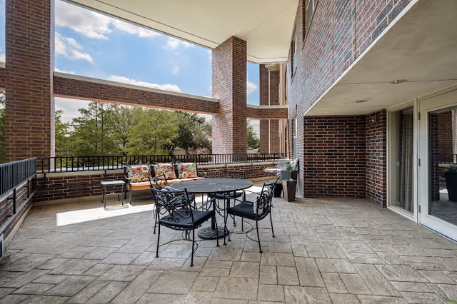 Outdoor covered patio area with a round metal table and four matching chairs. There is a cushioned bench with floral pillows along the brick wall railing. The patio overlooks green trees under a partly cloudy sky.