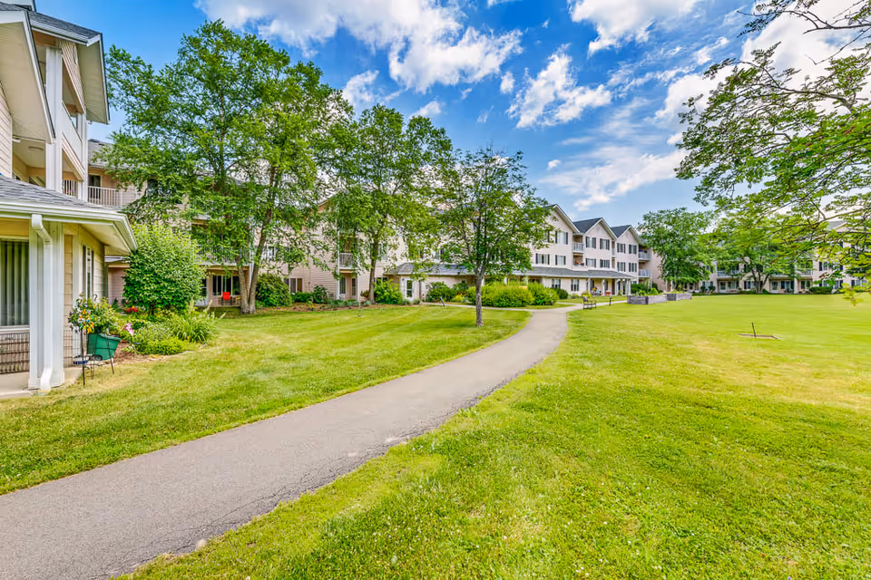 A wide paved pathway curves through a large, well-maintained grassy lawn with several trees. Surrounding the lawn are multi-story residential buildings with balconies, under a partly cloudy blue sky.