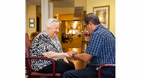 An elderly woman and a man sitting in chairs facing each other in a hallway or common area, holding hands and engaging in a warm, supportive interaction.