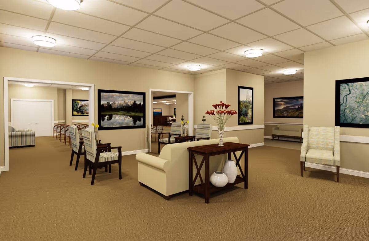 A spacious senior living facility common area with beige walls and carpet. The room is furnished with several cushioned chairs and sofas arranged around wooden tables. The tables hold decorative vases and a flower arrangement. The walls are decorated with framed landscape photographs. The ceiling has recessed lighting fixtures.