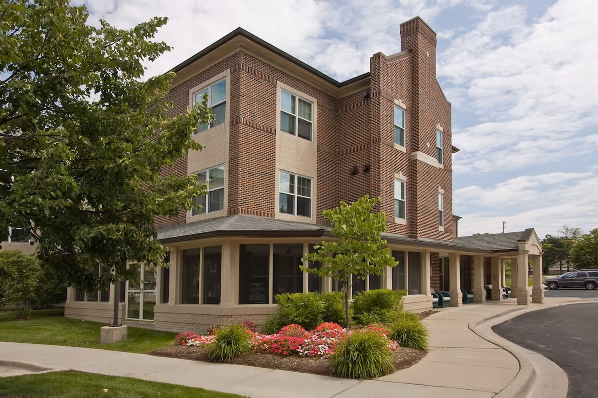 Exterior view of a three-story brick building with beige accents, surrounded by green trees and colorful flower beds, with a curved driveway and sidewalk in front.