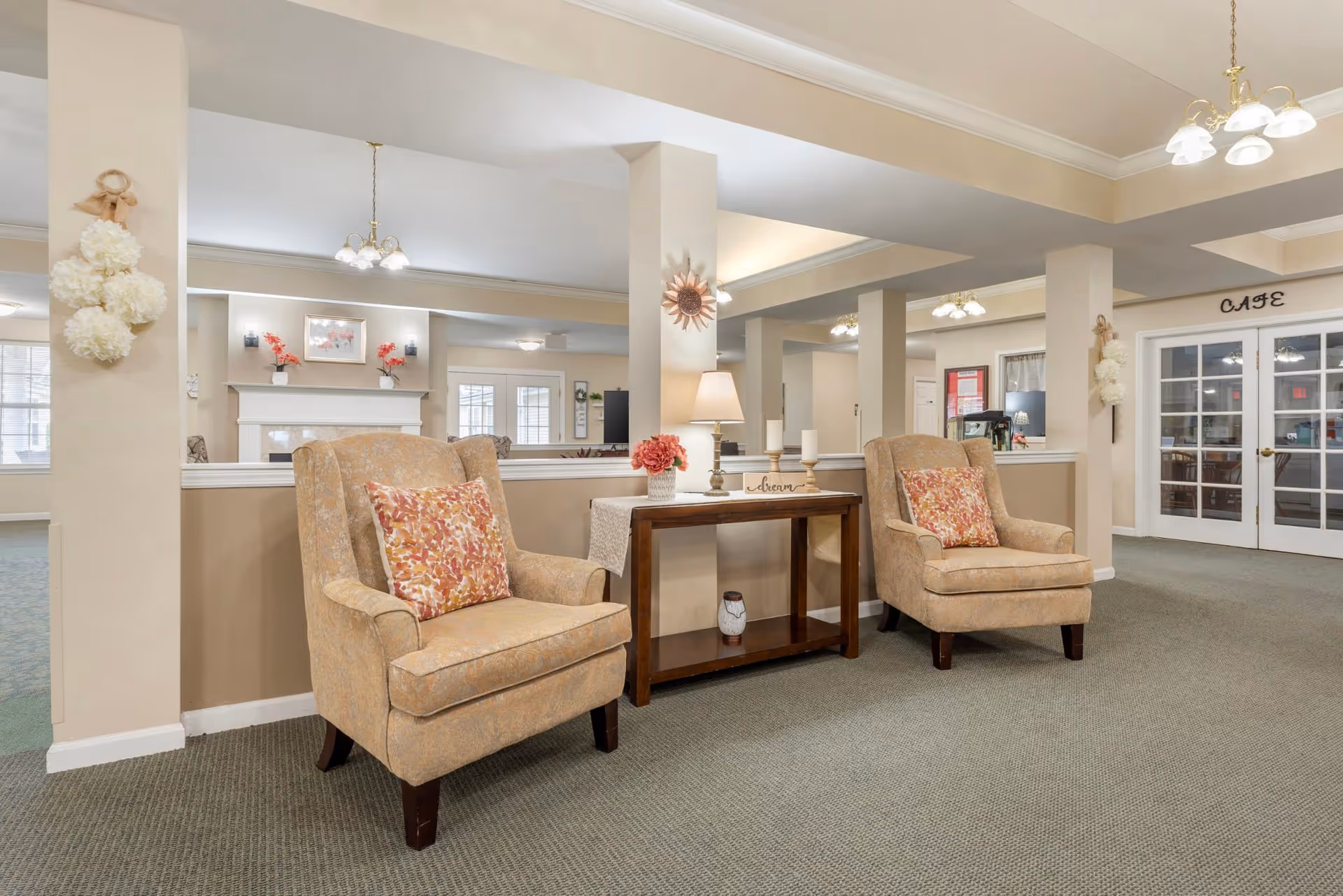 A cozy seating area in a senior living facility featuring two beige armchairs with floral cushions, a wooden console table with a lamp, candles, and a small flower arrangement. The background shows a fireplace, decorative wall hangings, and double doors labeled 'CAFE'. The space is well-lit with ceiling lights and has a neutral color palette.