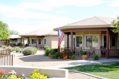 Exterior view of a single-story assisted living facility with beige walls and brown trim. The building has covered porches with seating areas and American flags displayed. There are potted plants and landscaped gravel areas with some greenery and trees.