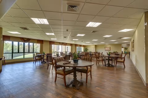 A spacious dining room with multiple round wooden tables and chairs arranged neatly. Large windows on one side allow natural light to fill the room, showing a view of greenery outside. The ceiling has recessed lighting and the floor is wooden. The room has a warm and inviting atmosphere.