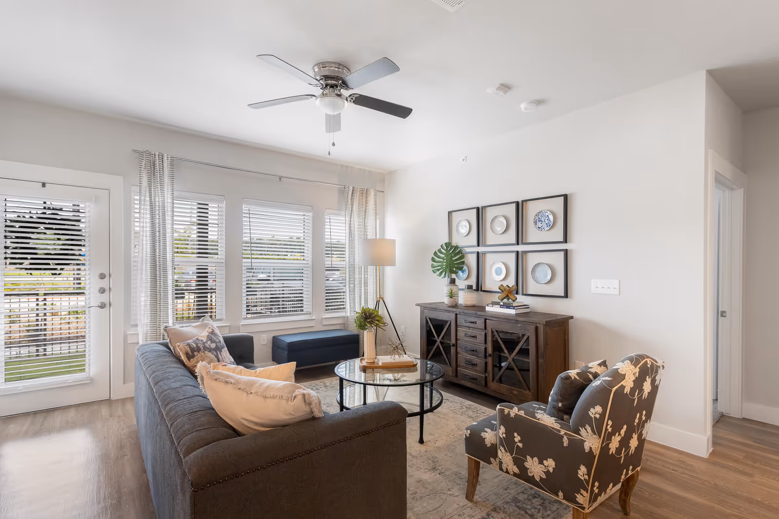 A modern living room featuring a gray sofa with decorative pillows, a glass coffee table, and a stylish armchair. The room has large windows with blinds, a decorative plant, and framed plates on the wall.