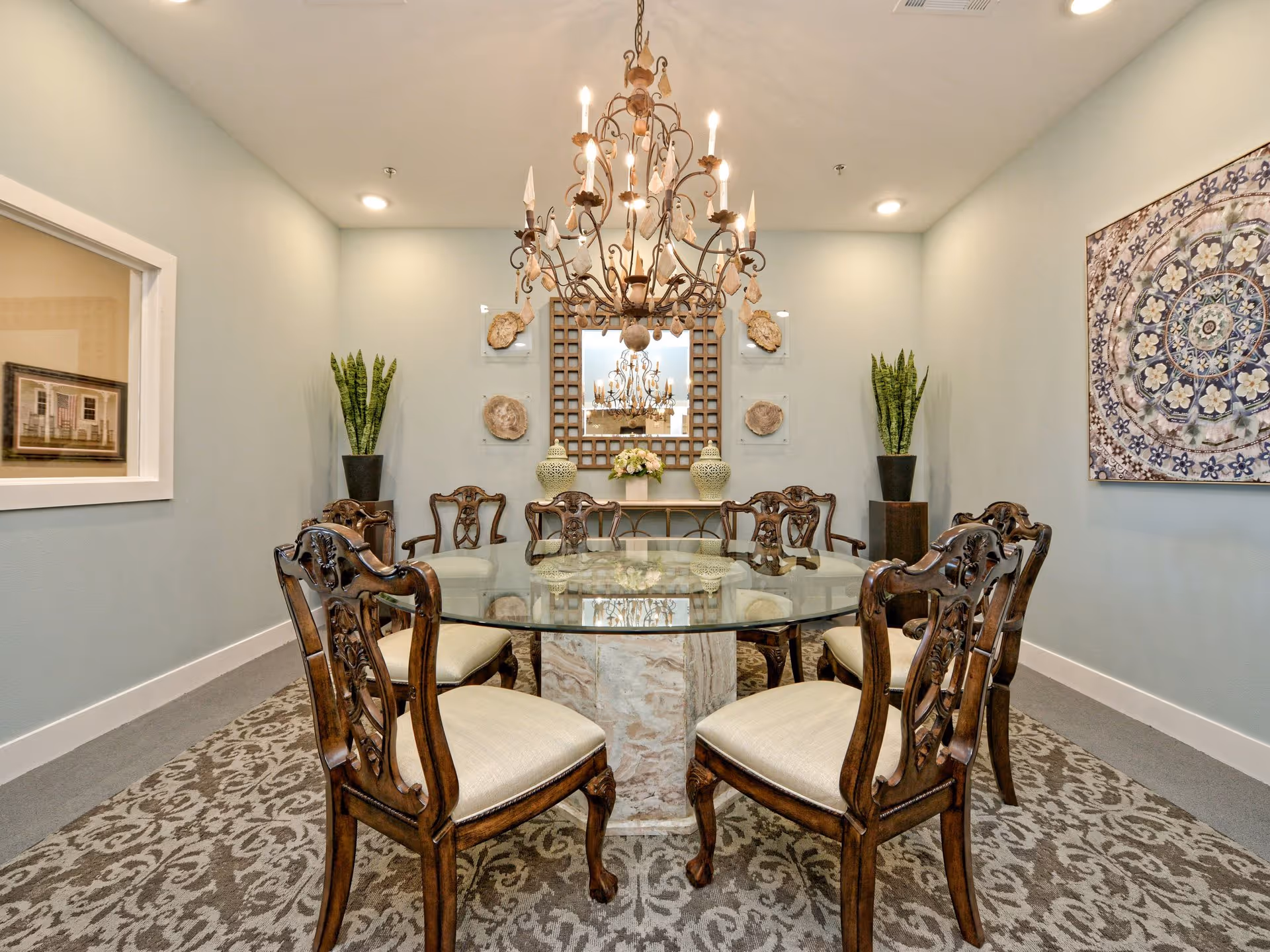 A formal dining room with a round glass table surrounded by eight ornate wooden chairs with cream cushions. A large decorative chandelier hangs above the table. The room features a patterned carpet, two tall potted plants in the corners, a large decorative wall mirror, and a framed artwork on the right wall.