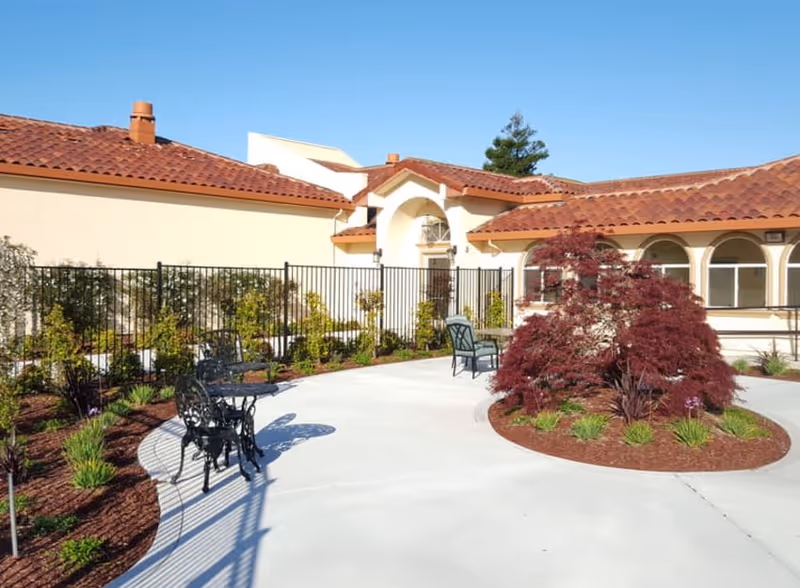 Outdoor courtyard area of a senior living facility with a paved walkway, black metal chairs and tables, landscaped garden beds with shrubs and a red-leafed tree, and a building with a tiled roof and arched windows in the background under a clear blue sky.