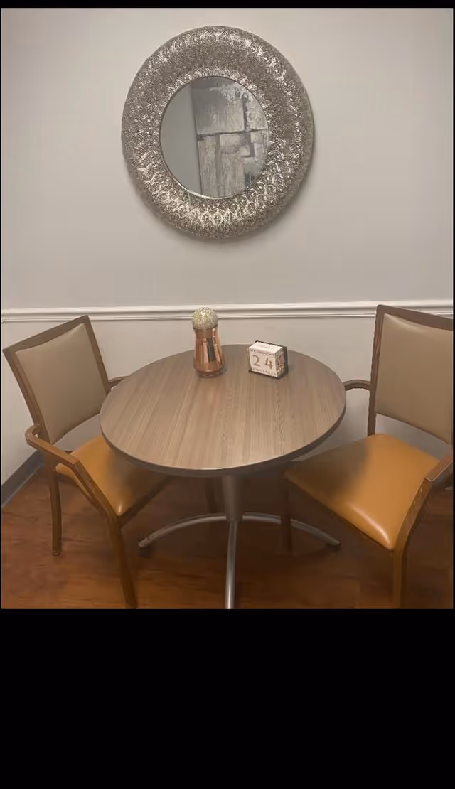 A small round wooden table with two chairs featuring tan cushions and wooden frames. On the table, there is a decorative item and a block calendar showing the date 24. Above the table, a round ornate mirror hangs on a plain light-colored wall with a white chair rail.