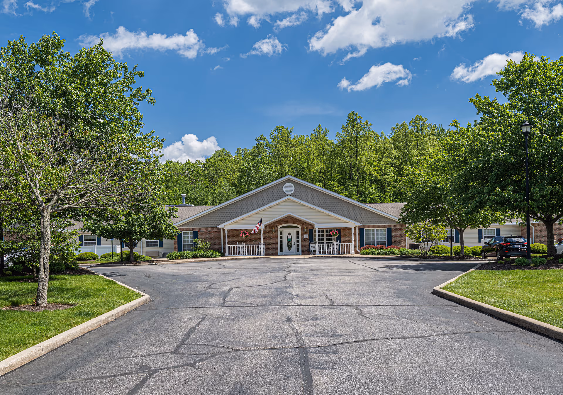 Front entrance of a single-story brick senior living building with a driveway, American flag, rocking chairs, and trees under a blue sky.