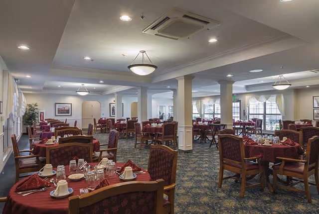 A spacious dining room in a senior living facility with multiple round tables covered with red tablecloths. Each table is set with cups, glasses, plates, and silverware. The room has carpeted floors, large windows with curtains, and ceiling lights providing bright illumination.