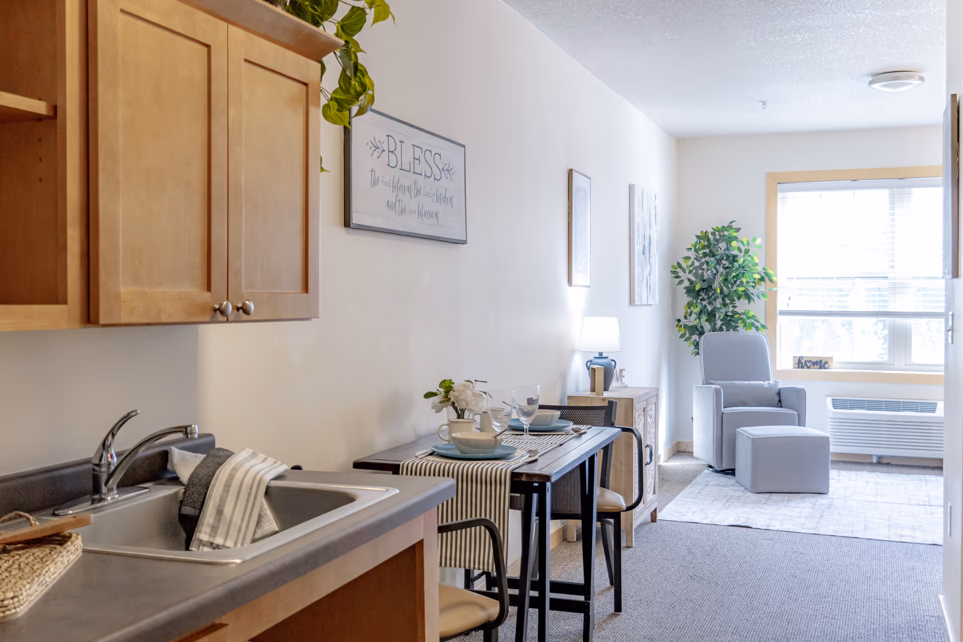 A bright and cozy interior space featuring a small kitchen area with a sink and wooden cabinets on the left. Adjacent to the kitchen is a dining table set for two with plates, bowls, and glasses. Further in the background is a living area with a comfortable gray armchair and matching ottoman, a side table with a lamp, a potted plant, and a large window letting in natural light. Wall decorations include framed artwork and a sign that reads 'BLESS'.