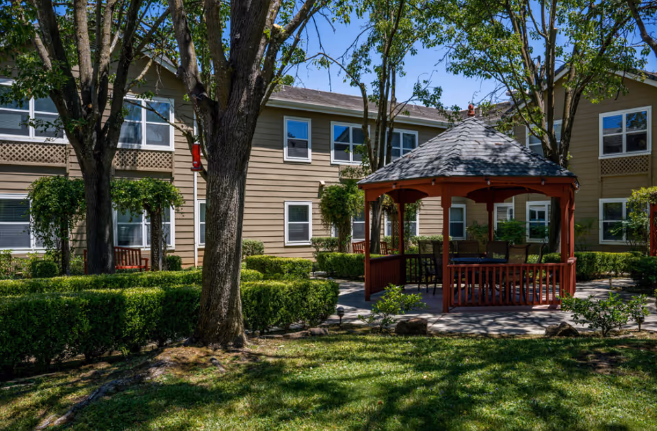 Outdoor garden area at a senior living facility with a wooden gazebo surrounded by trees, bushes, and a well-maintained lawn. The building with multiple windows is visible in the background under a clear blue sky.