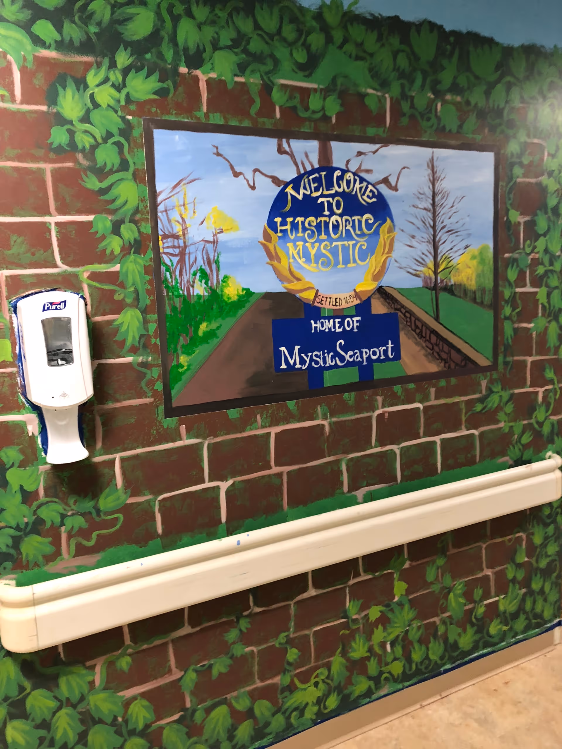 A painted mural on a brick wall depicting a sign that reads 'Welcome to Historic Mystic, Home of Mystic Seaport' with greenery and trees in the background. There is a white Purell hand sanitizer dispenser mounted on the wall to the left and a beige handrail running horizontally below the mural.