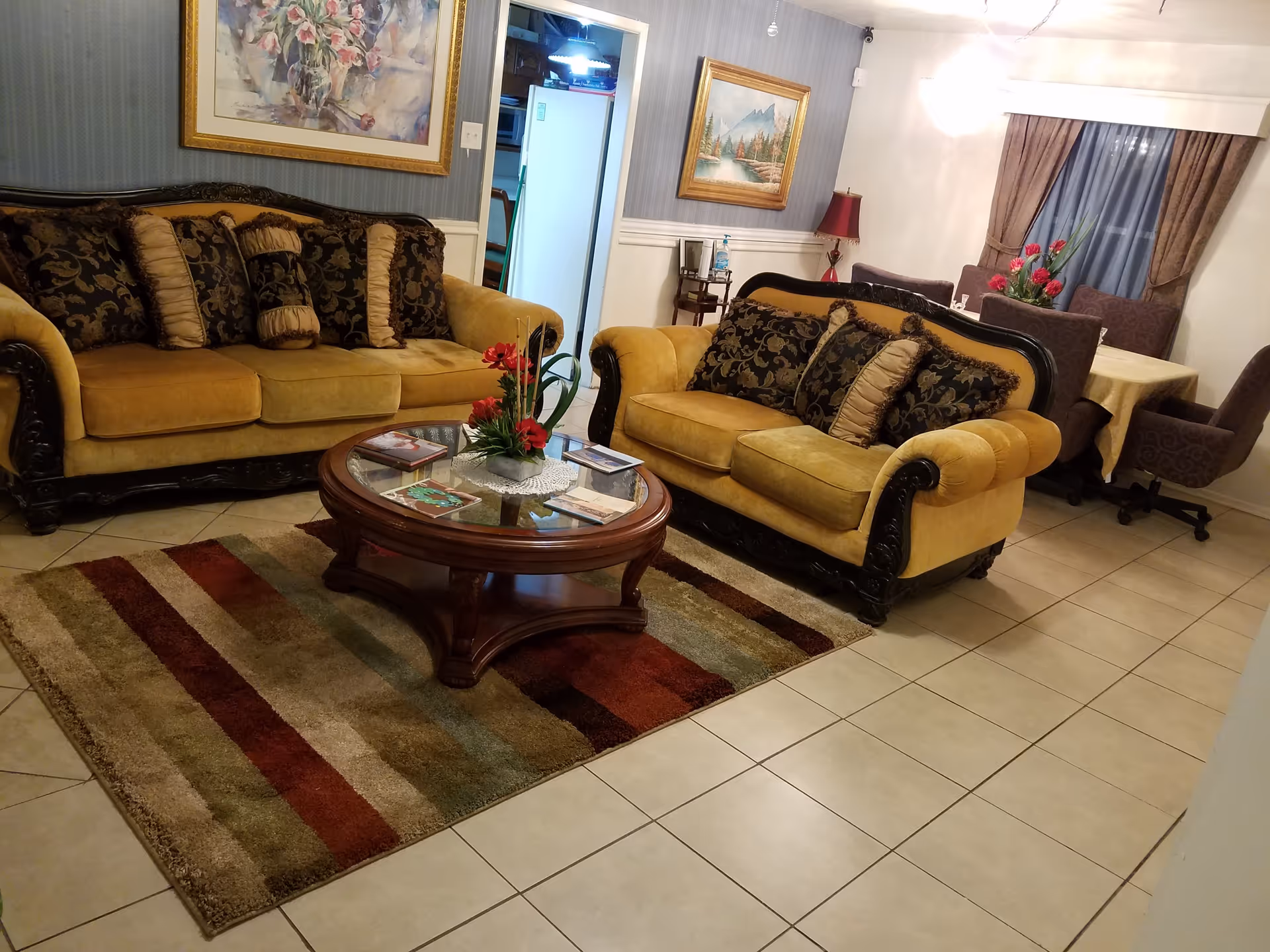 A cozy living room with two mustard yellow sofas adorned with patterned cushions, a wooden coffee table with a glass top and floral centerpiece, a striped area rug, and framed paintings on the walls. In the background, there is a dining area with a table, chairs, and a window with curtains.