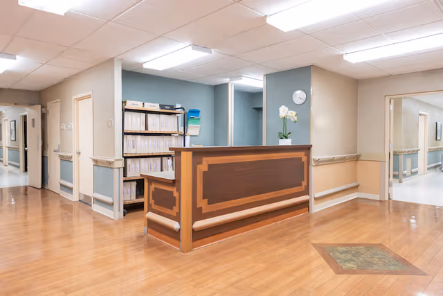 A clean and well-lit reception area in a healthcare facility with a wooden reception desk, shelves filled with files, a clock on the wall, and a potted orchid plant on the desk. The floor is wooden, and there are hallways extending to the left and right.