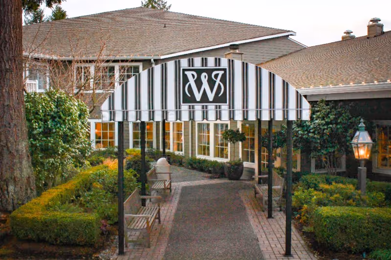 Entrance pathway to a building with a black and white striped canopy featuring a stylized 'W' logo. The pathway is lined with benches and greenery, including trimmed bushes and potted plants. The building has multiple windows and a shingled roof.