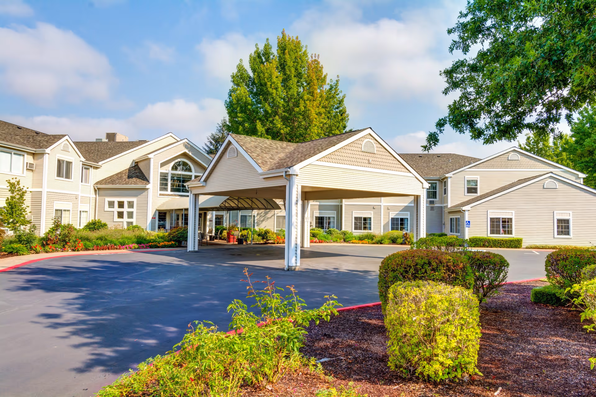 Exterior view of The Oaks at Lebanon senior living facility showing a large covered entrance with beige siding, multiple windows, landscaped bushes, and trees under a partly cloudy sky.