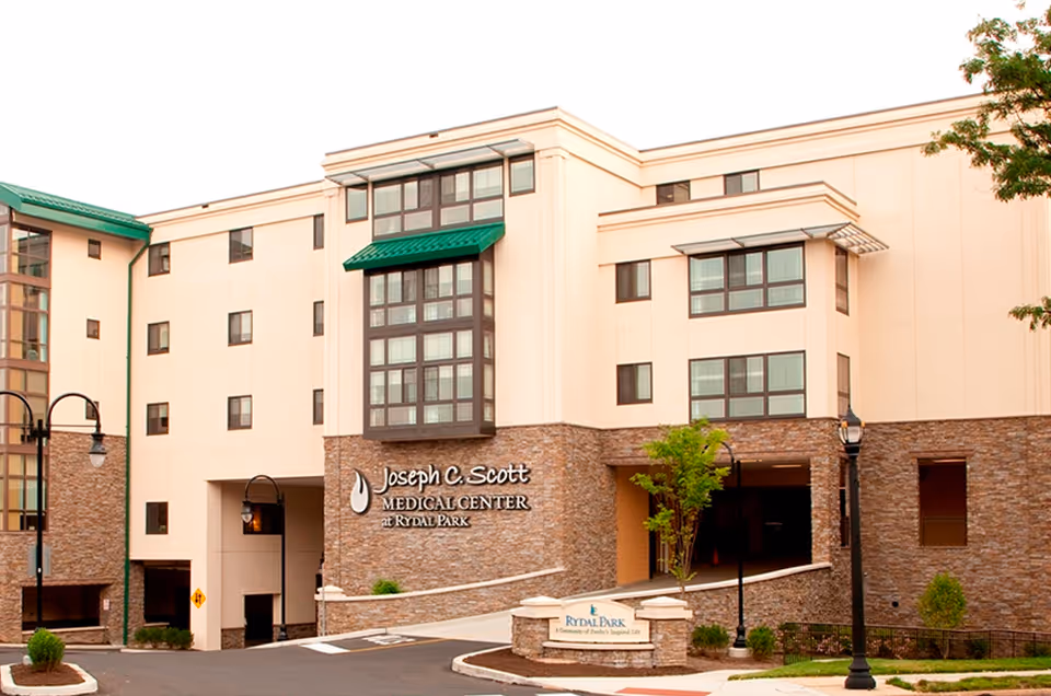 Exterior view of the Joseph C. Scott Medical Center at Rydal Park, a multi-story building with beige walls and stone accents, green window awnings, and a sign for Rydal Park at the entrance driveway.