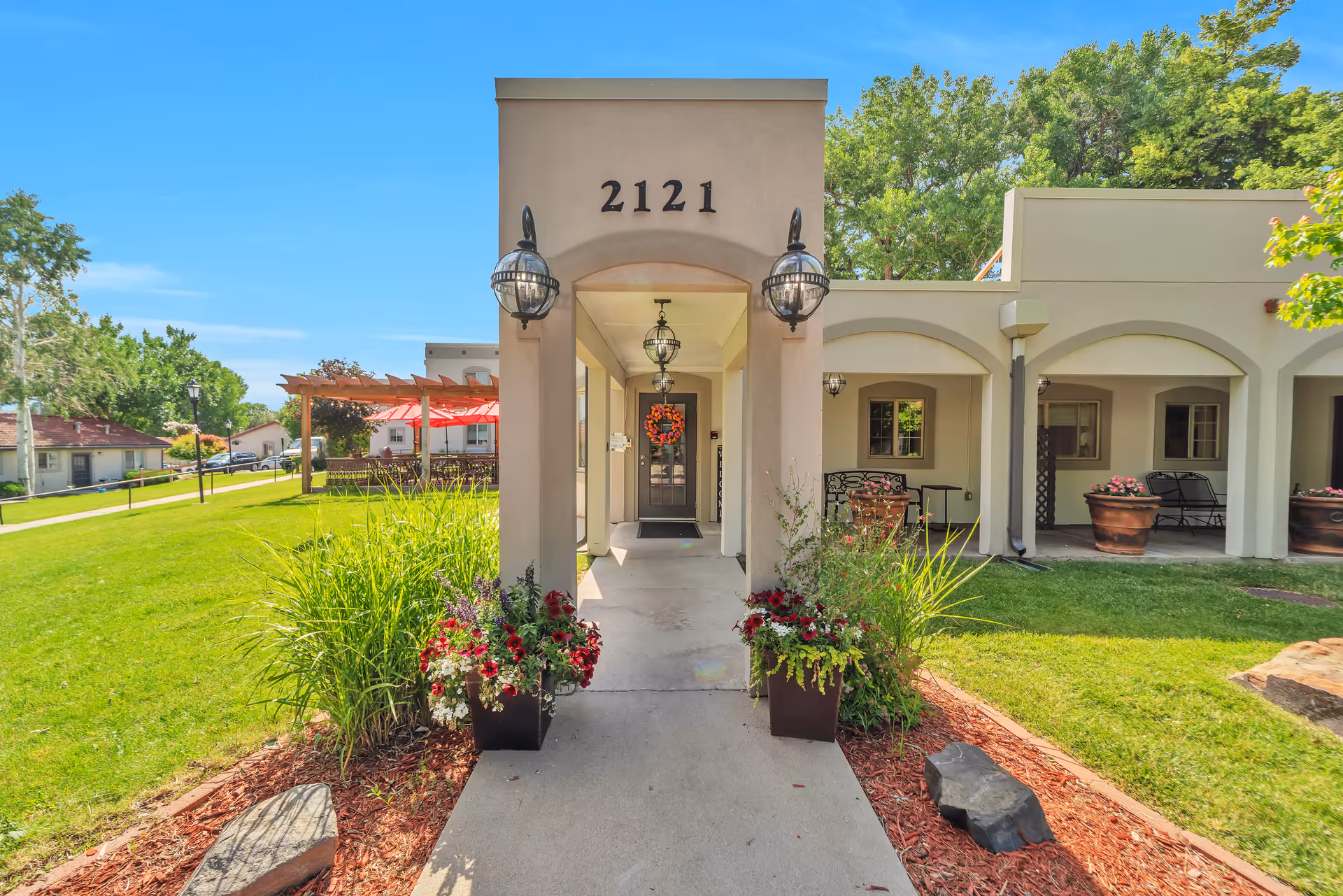 Front entrance of a senior living facility with the number 2121 displayed above the doorway. The entrance is framed by two large lantern-style lights and flanked by planters with flowers and greenery. The building has a beige exterior with arches and a covered walkway. There is a green lawn and trees surrounding the building under a clear blue sky.