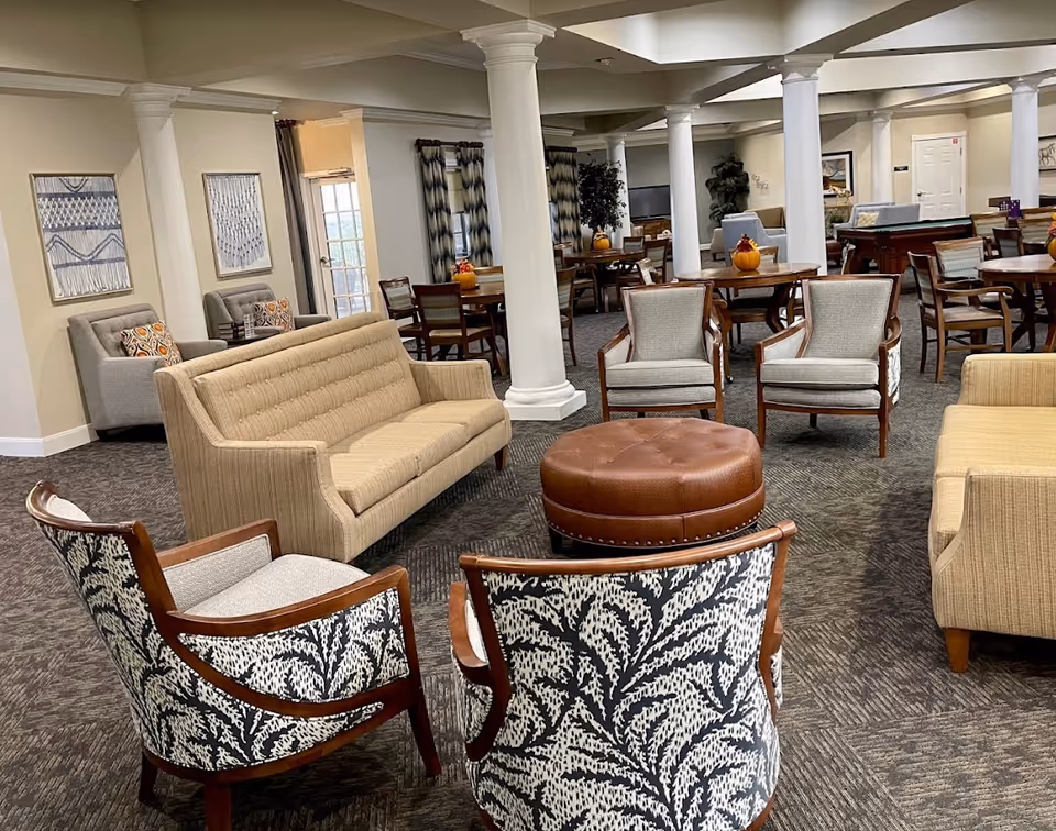 Open communal lounge in a senior living facility with sofas, armchairs, a round leather ottoman, dining tables and white columns.