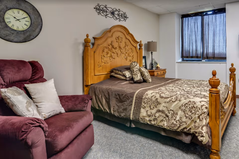 A bedroom with a wooden bed frame featuring intricate carvings, a bed with brown and beige patterned bedding, a wooden nightstand with a lamp and decorative items, a large maroon armchair with pillows, a wall clock, and a window with dark curtains.