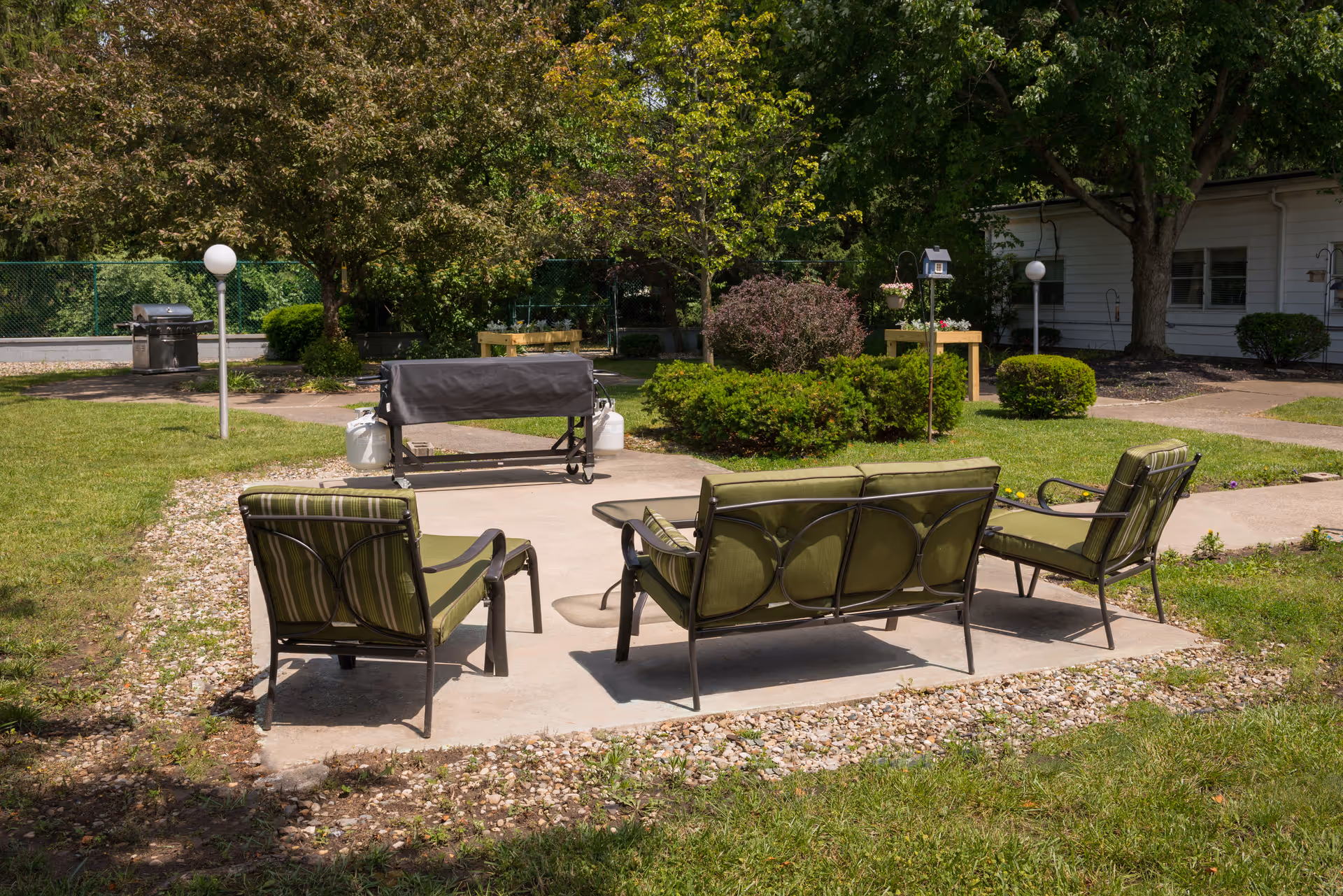 Outdoor patio area with green cushioned metal chairs and a loveseat arranged around a concrete slab. In the background, there is a covered grill, trees, bushes, and a white building partially visible. The area is surrounded by grass and a gravel border.