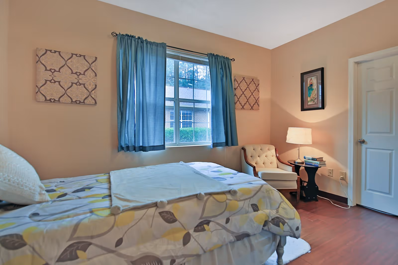 A tidy bedroom with a bed, a window with blue curtains, an upholstered chair and a small bedside table with a lamp.