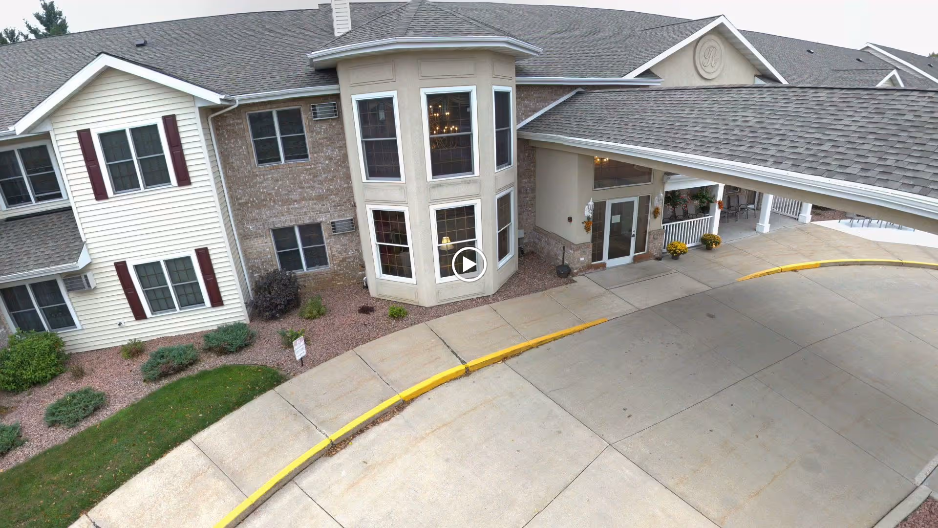 Exterior view of Renaissance by Rennes - Wisconsin Rapids showing the entrance with a covered drop-off area, large windows, and a combination of brick and siding on the building facade. Landscaping with bushes and a curved driveway are visible.