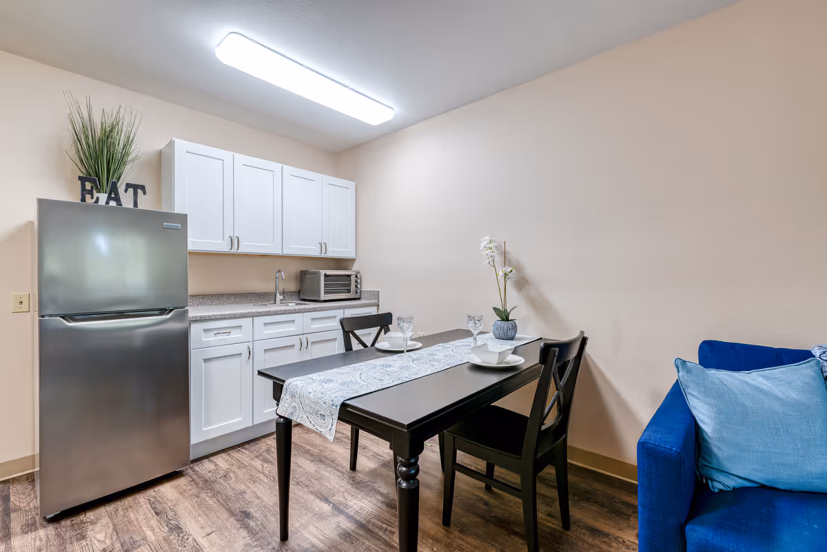 A small kitchen and dining area featuring white cabinets, a stainless steel refrigerator with a plant and 'EAT' letters on top, a countertop with a toaster oven, a dark wooden dining table set for two with a decorative runner and a small flower vase, and a blue couch with light blue pillows.