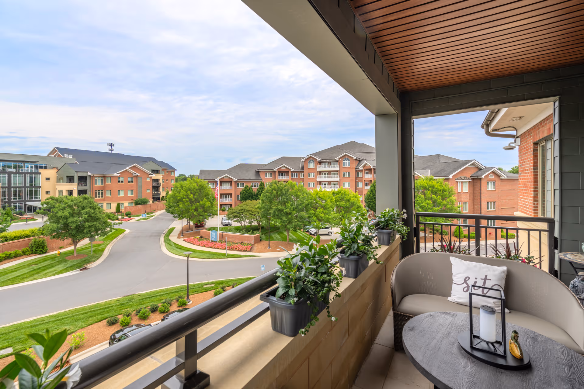 View from a covered balcony with a small round table, a cushioned curved bench with a pillow that says 'sit', and several potted plants on the balcony railing. The balcony overlooks a landscaped road with trees and multiple brick residential buildings under a partly cloudy sky.