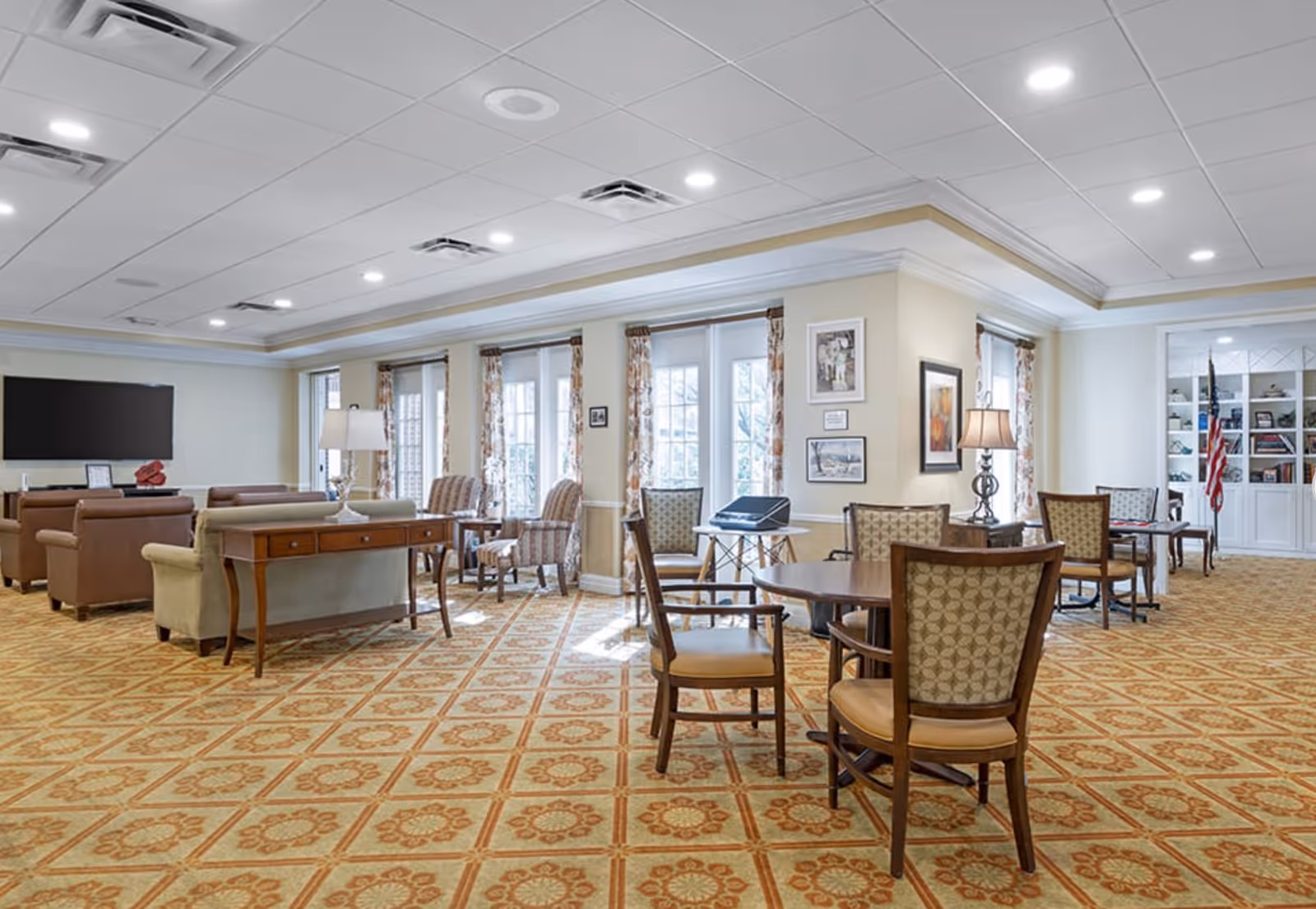 A spacious and well-lit common area in a senior living facility featuring patterned carpet, multiple seating arrangements including armchairs and wooden chairs around tables, a large flat-screen TV mounted on the wall, lamps, framed pictures on the walls, and large windows with floral curtains allowing natural light to enter.