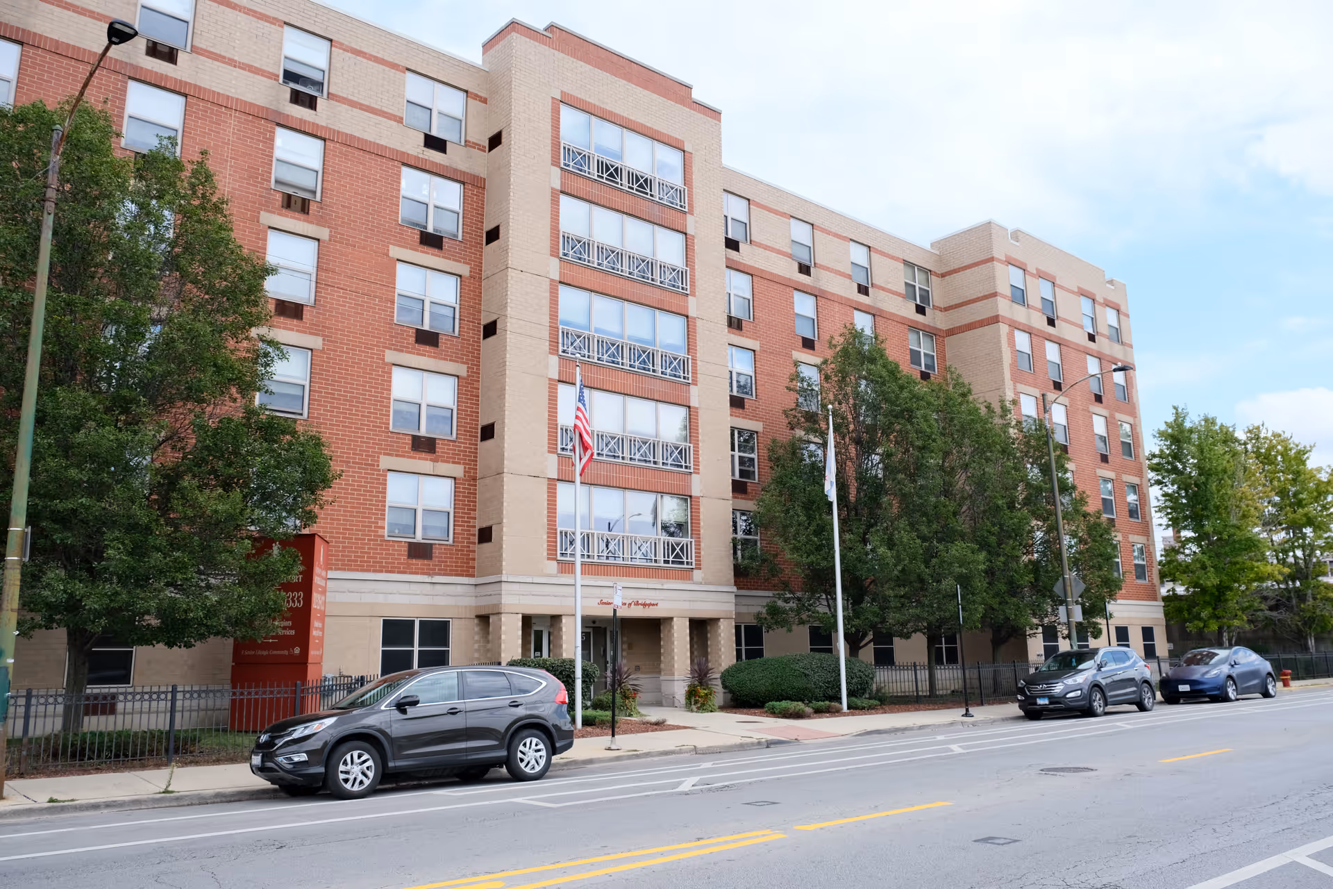 Exterior view of a multi-story senior living facility named Senior Suites of Bridgeport, featuring a brick and beige facade, several windows, two flagpoles with flags, and parked cars along the street in front.