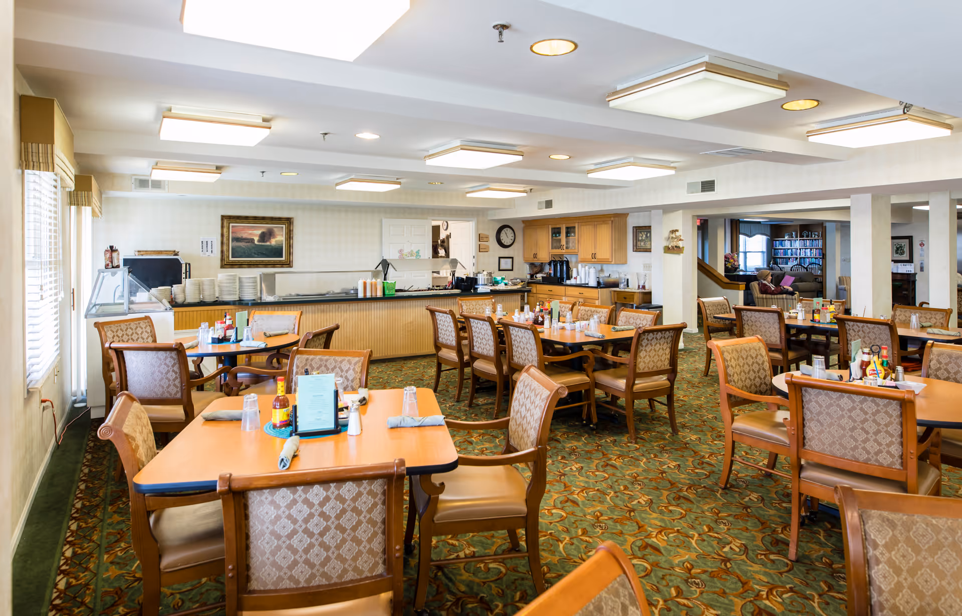 Bright communal dining room with multiple wooden tables and chairs, a serving counter at the back, and patterned carpet.
