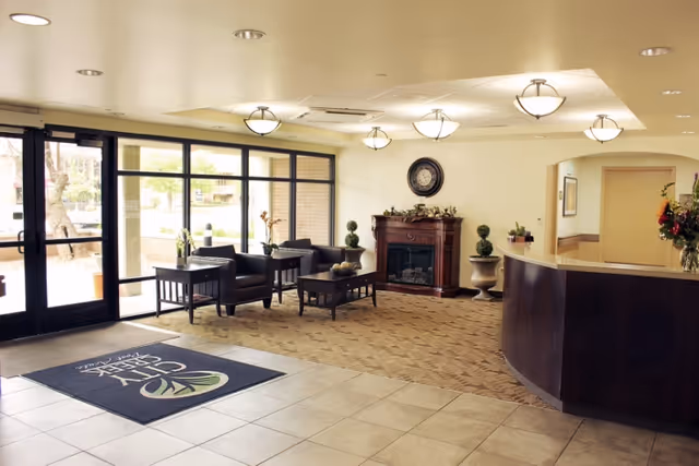 Lobby area of City Creek Post Acute facility with large glass entrance doors, a seating area with black chairs and tables, a decorative fireplace with a clock above it, and a curved reception desk with flowers on it.