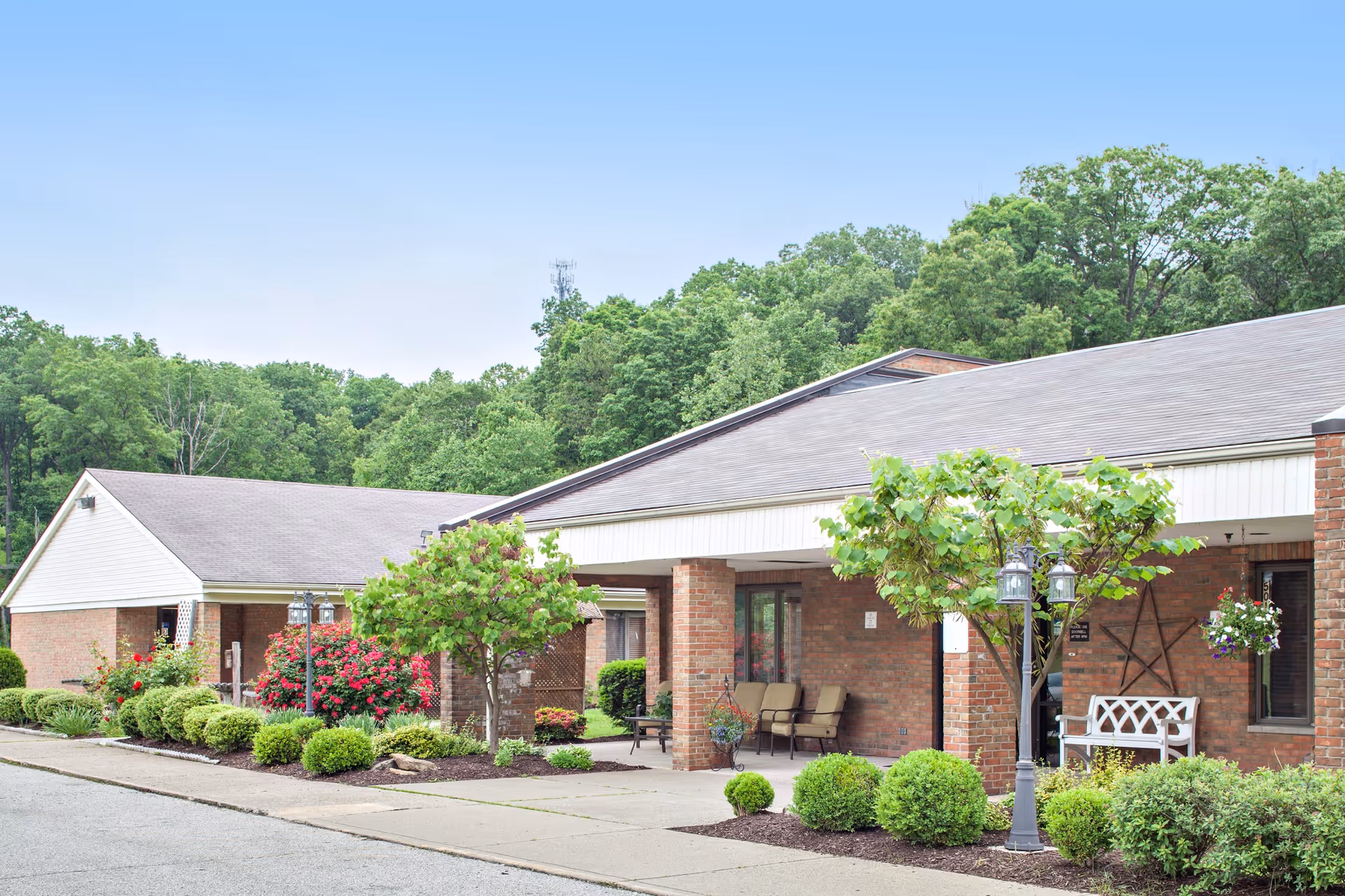 Exterior view of a single-story brick building with a covered entrance, surrounded by well-maintained landscaping including bushes, small trees, and flowering plants under a clear blue sky.