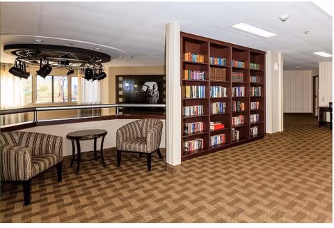 Interior view of a senior living facility lounge area with two striped armchairs and a small round table between them. A large bookshelf filled with books is positioned against a wall. The carpet has a geometric pattern, and there is a lighting fixture with spotlights hanging from the ceiling. Windows with curtains are visible in the background.