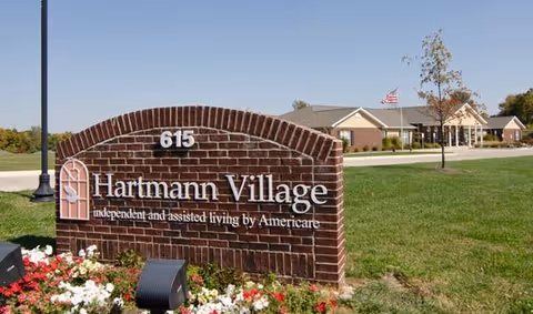 Brick sign for Hartmann Village independent and assisted living by Americare with the number 615, set in a landscaped area with flowers and grass. In the background, there is a single-story building with an American flag on a flagpole and a clear blue sky.