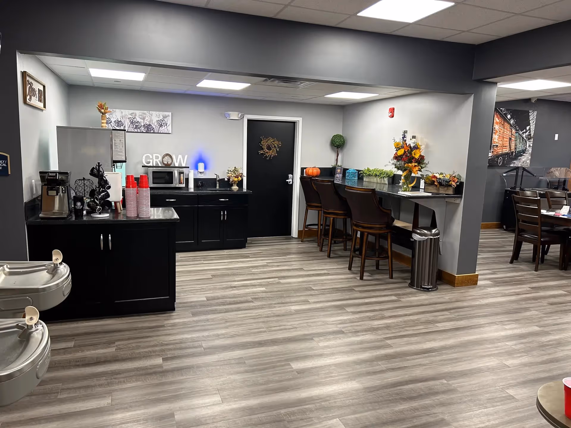 Interior view of a common area in a senior living facility featuring a coffee station with a coffee maker, microwave, and red disposable cups on black cabinets. There is a counter with three high chairs decorated with fall-themed flowers and a small pumpkin. The floor is light wood laminate, and the walls are painted gray. In the background, there is a dining area with tables and chairs and a large photo of a train on the wall.