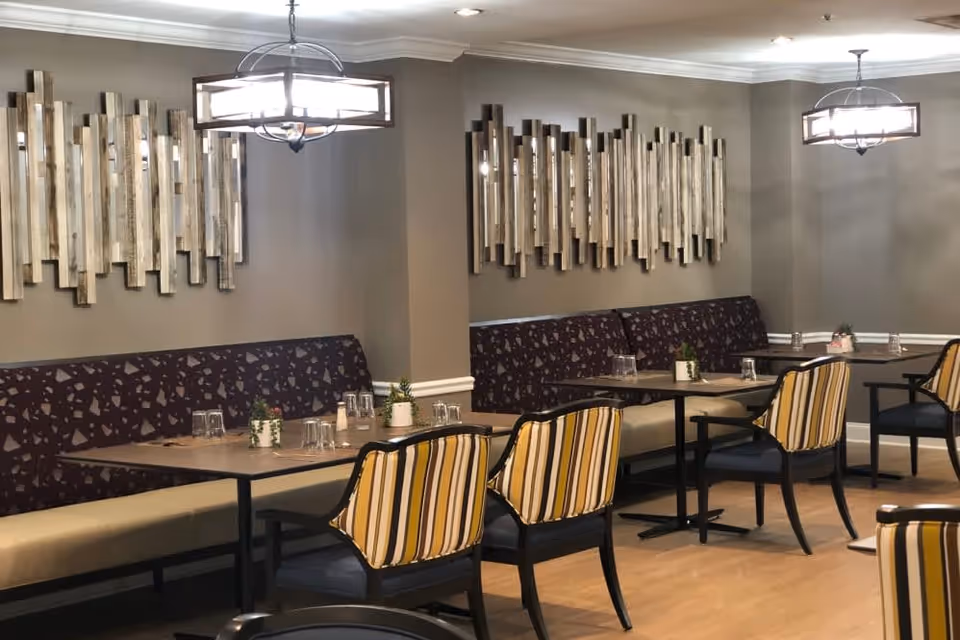 Dining area with tables and chairs, featuring striped cushioned chairs and a long bench with patterned upholstery. The walls are decorated with vertical wooden art pieces, and two modern chandeliers hang from the ceiling.