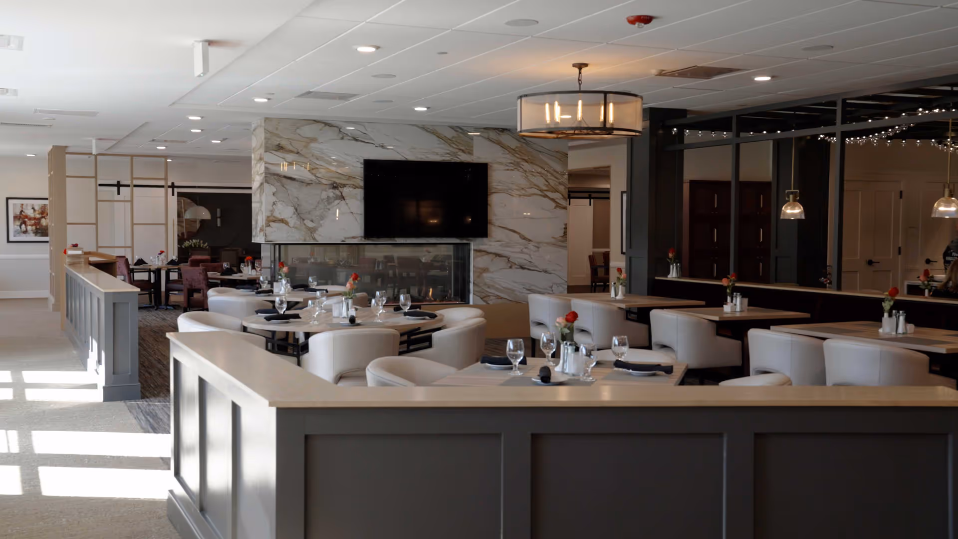 A bright, modern dining room with set tables and cream upholstered chairs, a marble-clad fireplace wall with a mounted TV, and overhead lighting.